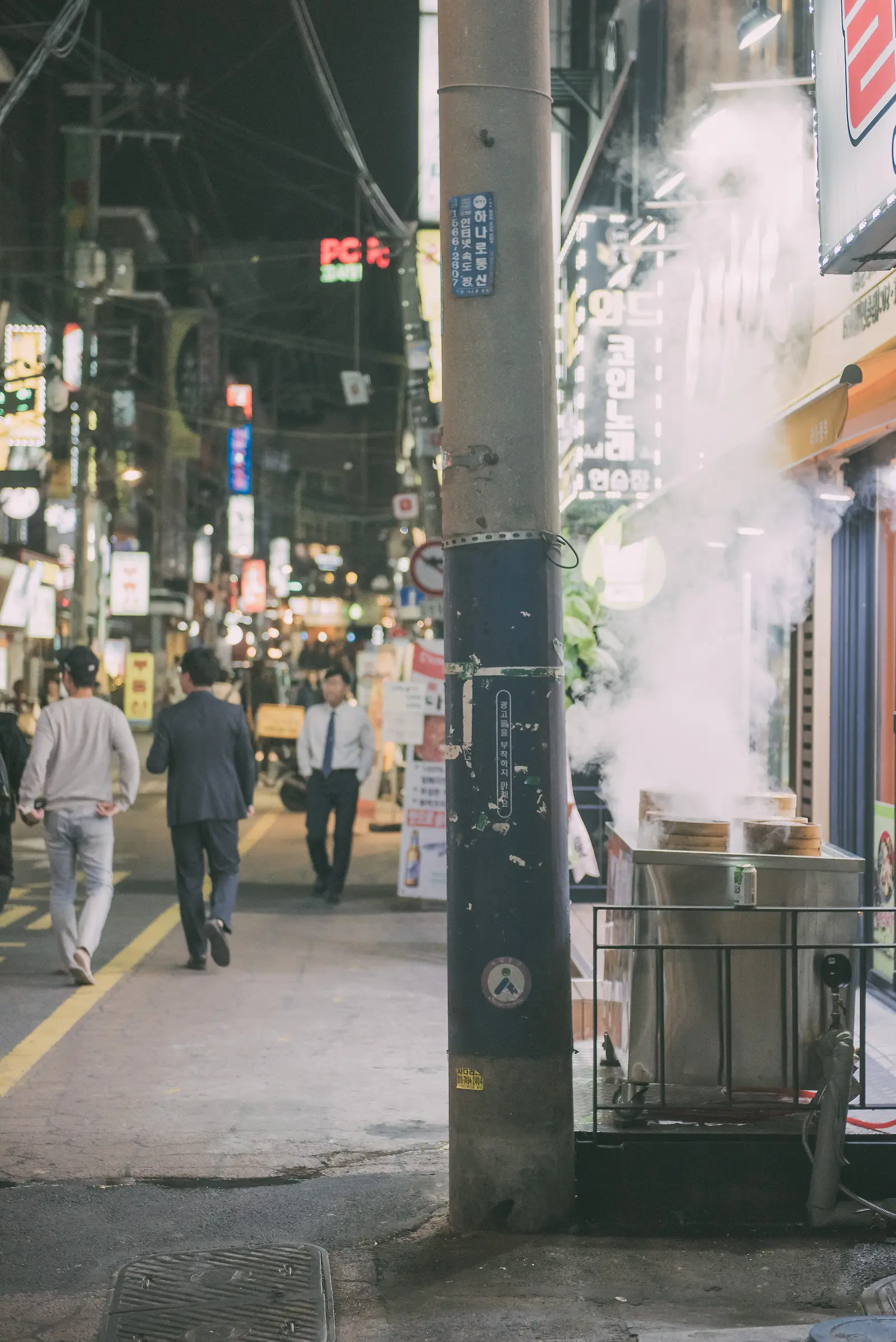 밤거리에서 음식점 앞 김이 오르는 골목을 걷는 사람들