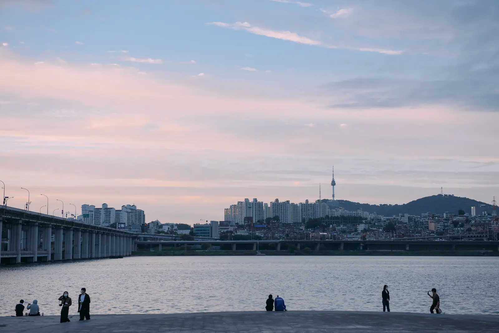 분홍빛 노을 아래 강변과 건물들, 물가를 걷는 사람들