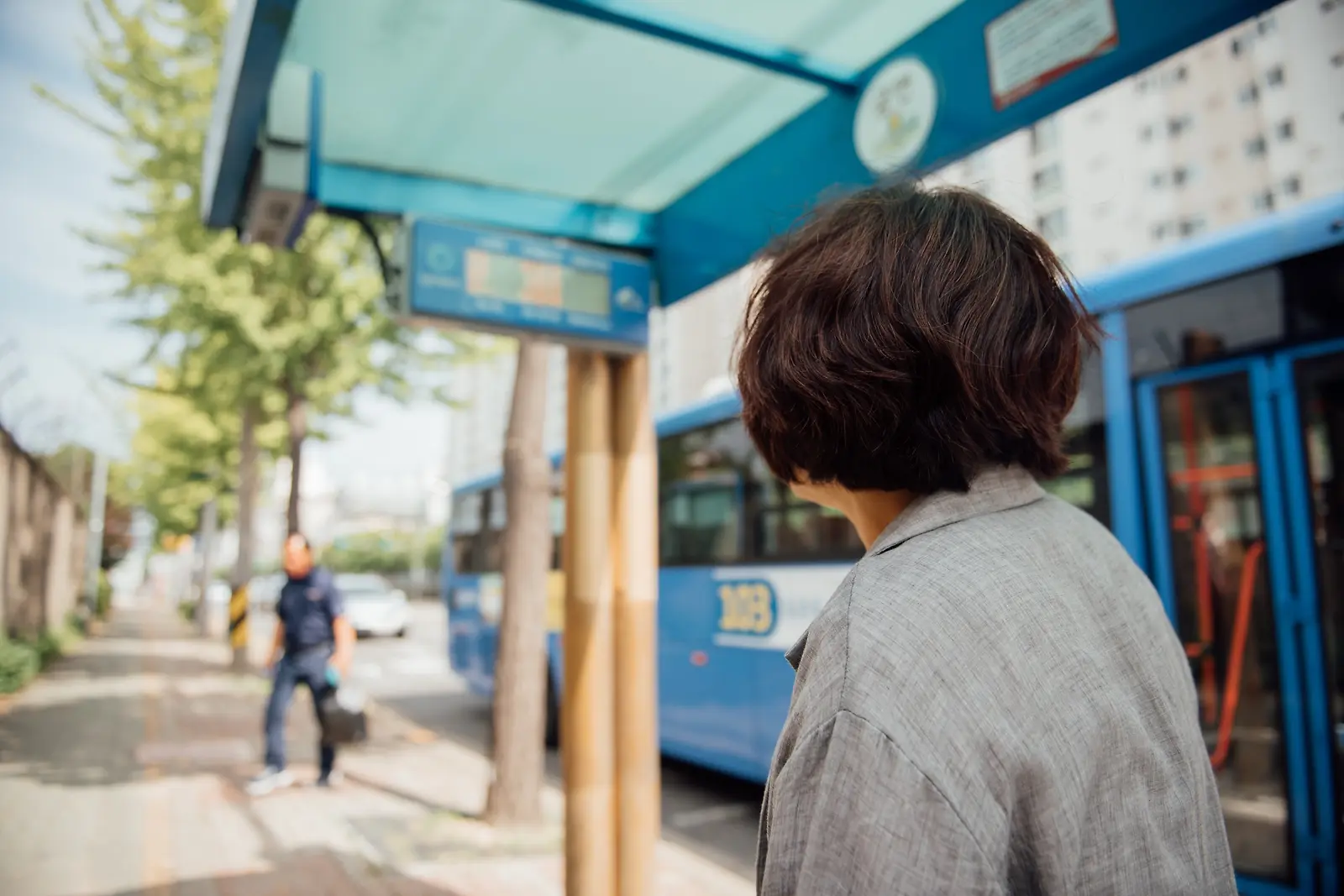 버스 정류장 안내판을 바라보며 기다리는 어머니의 뒷모습